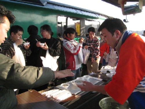 緑色の列車が背景に見える駅のホームで、法被を着た男性が観光客たちに温かい汁物が入った器を手渡しているセレモニーの様子の写真