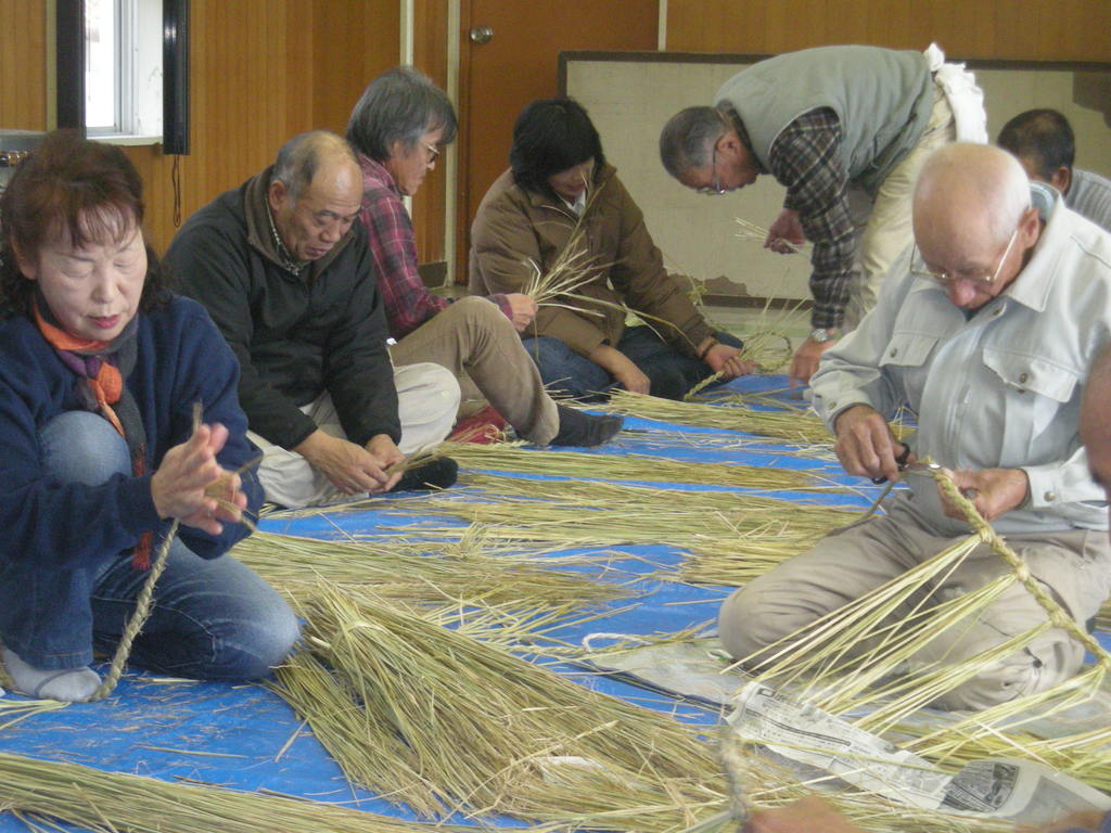ブルーシートを敷いた屋内床で、数人の参加者が集まり、藁を使ってしめ縄を編んでいる様子の写真