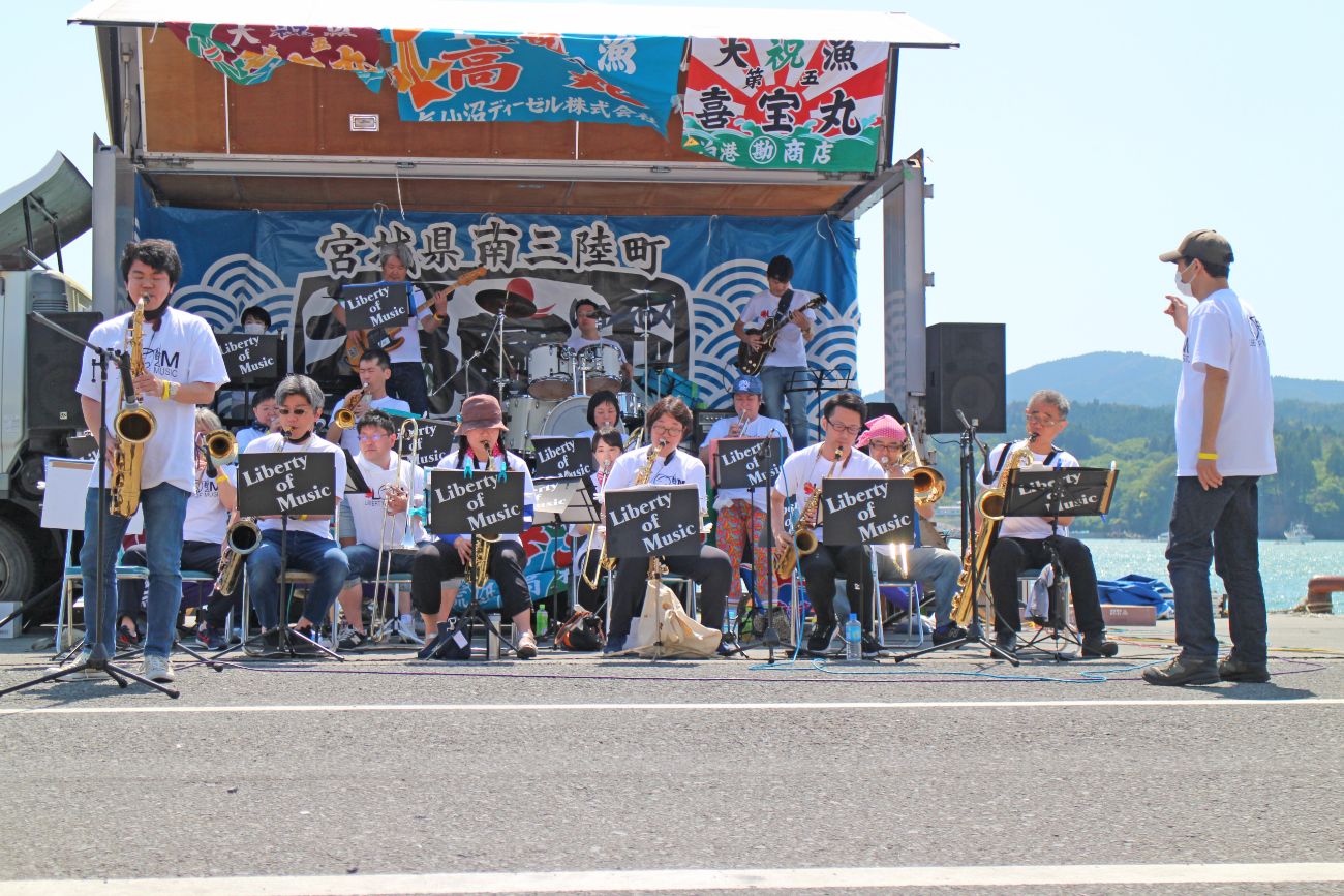 「宮城県南三陸町」の横断幕が掲げられた海辺の特設ステージで「Liberty of Music」と書かれた譜面台の前でサックスやトランペット、ドラム、ギターなどの楽器を担当するメンバーが演奏しており、左前でサックスを持ちソロパートを演奏する男性メンバーと右端で指揮をとる男性が立っている様子が演奏中の写真
