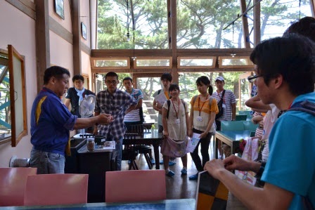 木の窓枠から外の緑が見える室内で、男性がテーブル越しに右手に持った物を見せながら説明をし、それを若者たちが囲んで聞いている写真