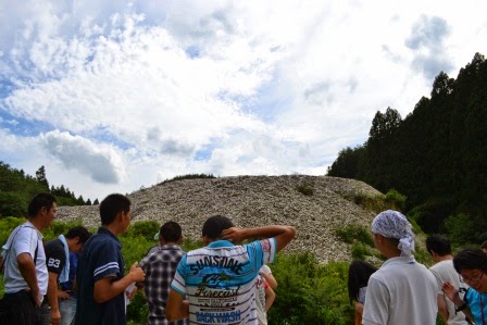 森の中の空き地に積み上がった大量の貝殻の山を、十数人の人たちが観察している写真