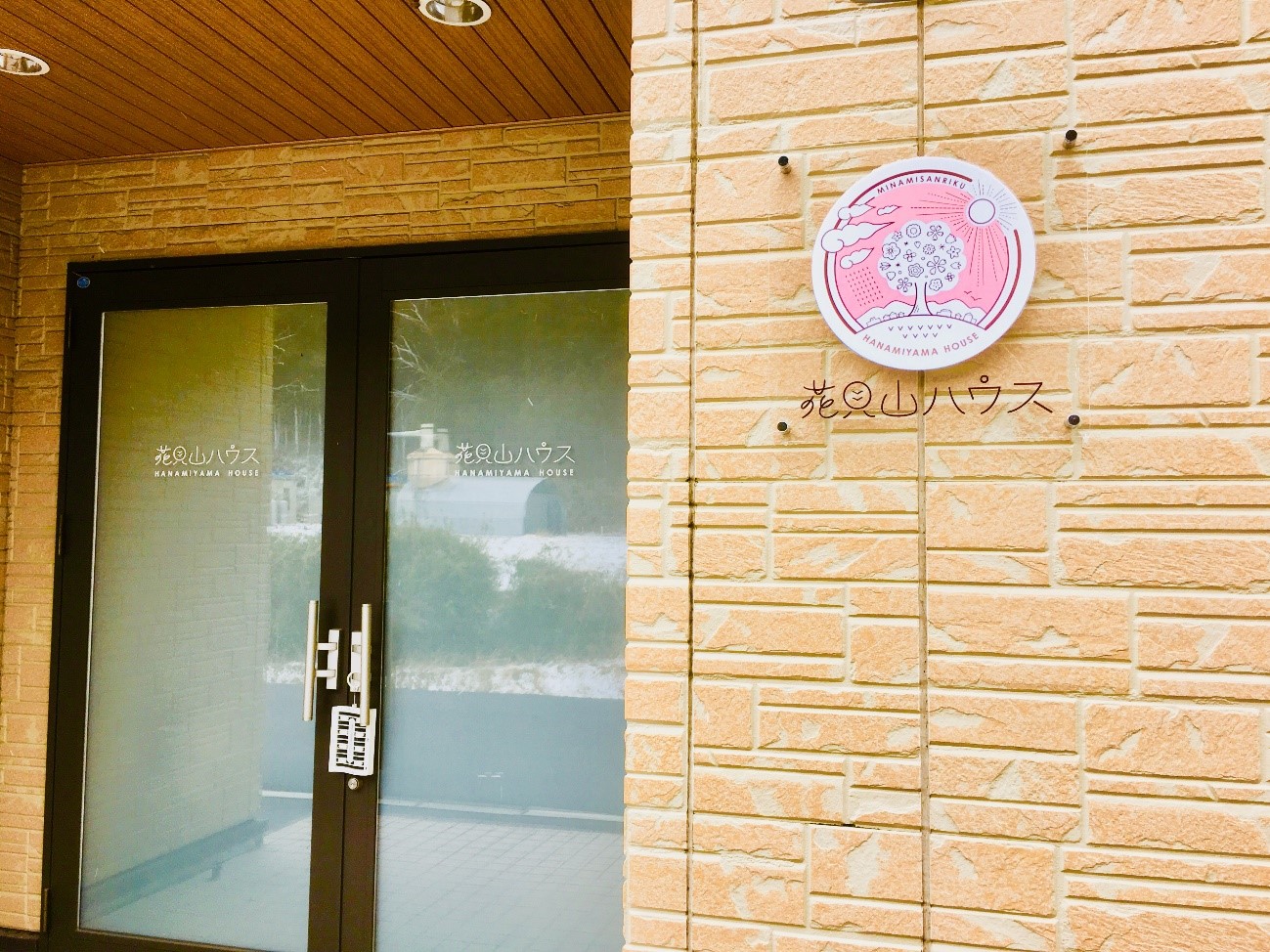 「花見山ハウス」と書かれたガラス扉と、右側のベージュ色の壁に丸いロゴマークの看板が取り付けられている玄関の写真