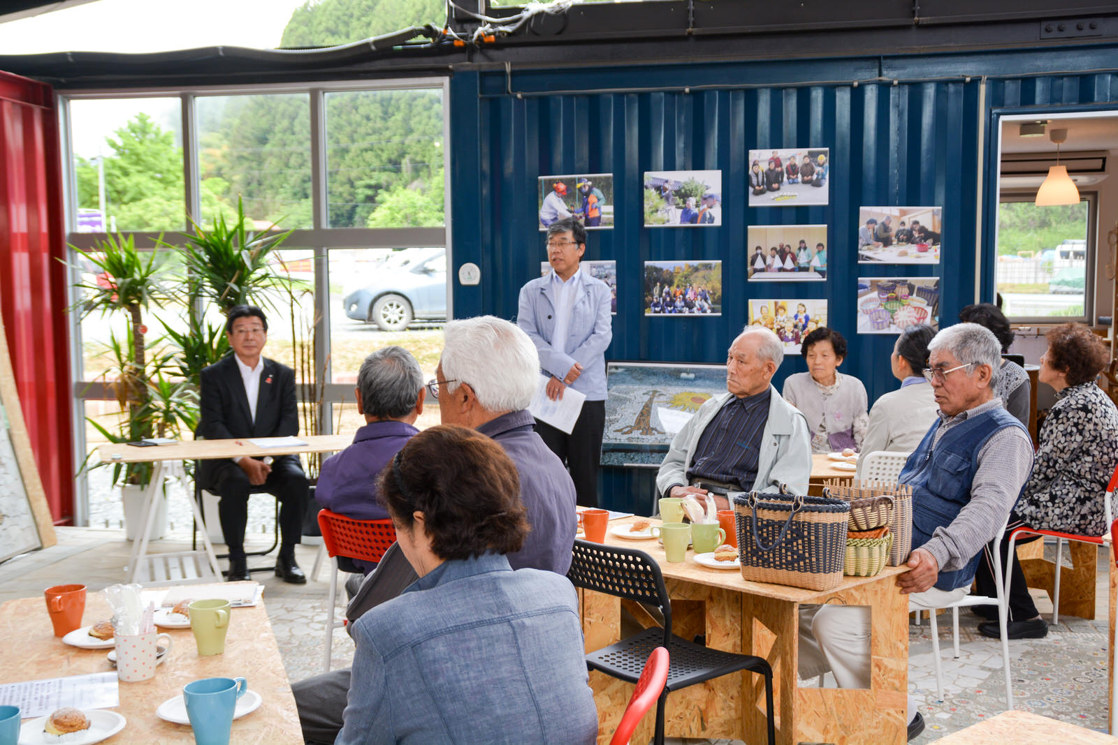 窓を背にして席に座った町長の横で、複数の写真が貼られた青い壁の前に資料を手にした男性が立っており、その前では木製テーブルを囲んでたくさんの高齢者の方々が座っている写真
