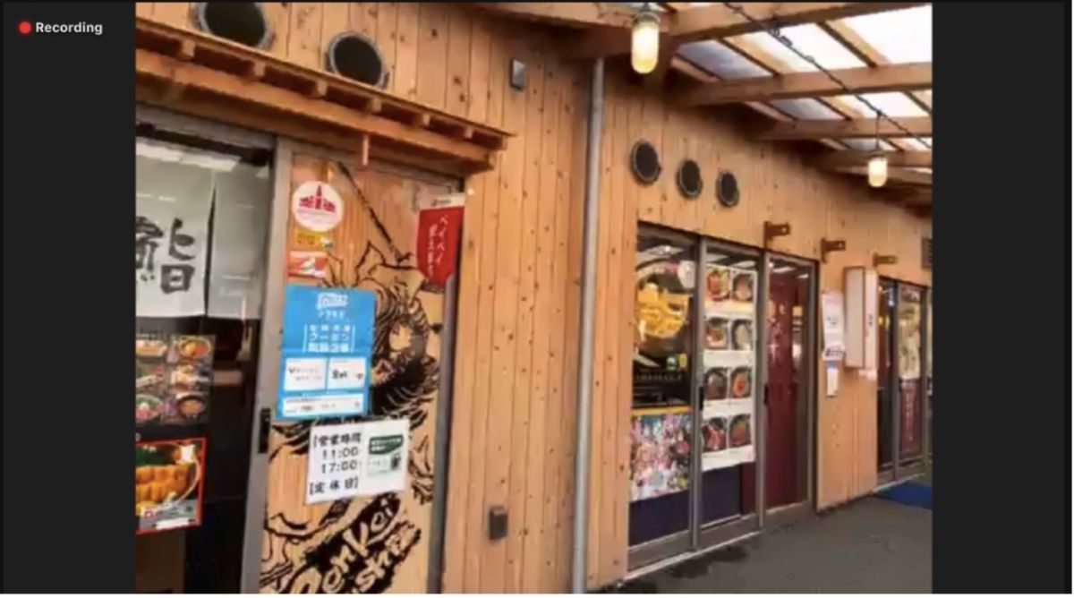 ドアにはメニューや営業情報のポスターが多数貼られた、木の外壁に囲まれた飲食店の並びが写っているスクリーンショット画像