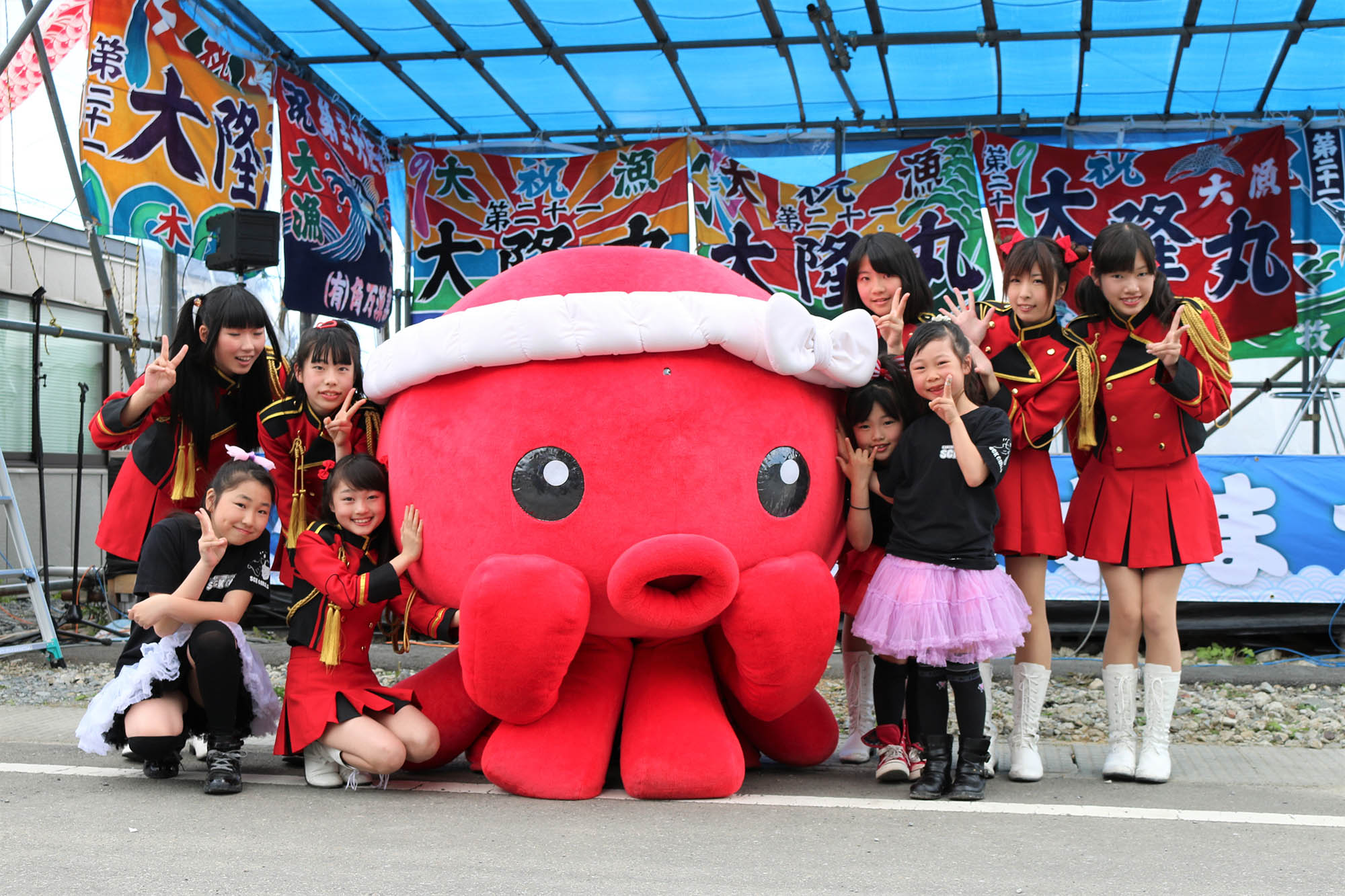 赤を基調に黒のアクセントがある衣装を着た子どもたちと黒い服にピンクのスカートを履いた子どもたちが、オクトパス君の周りで笑顔で手を挙げてポーズをとっている、屋外のイベントステージの写真