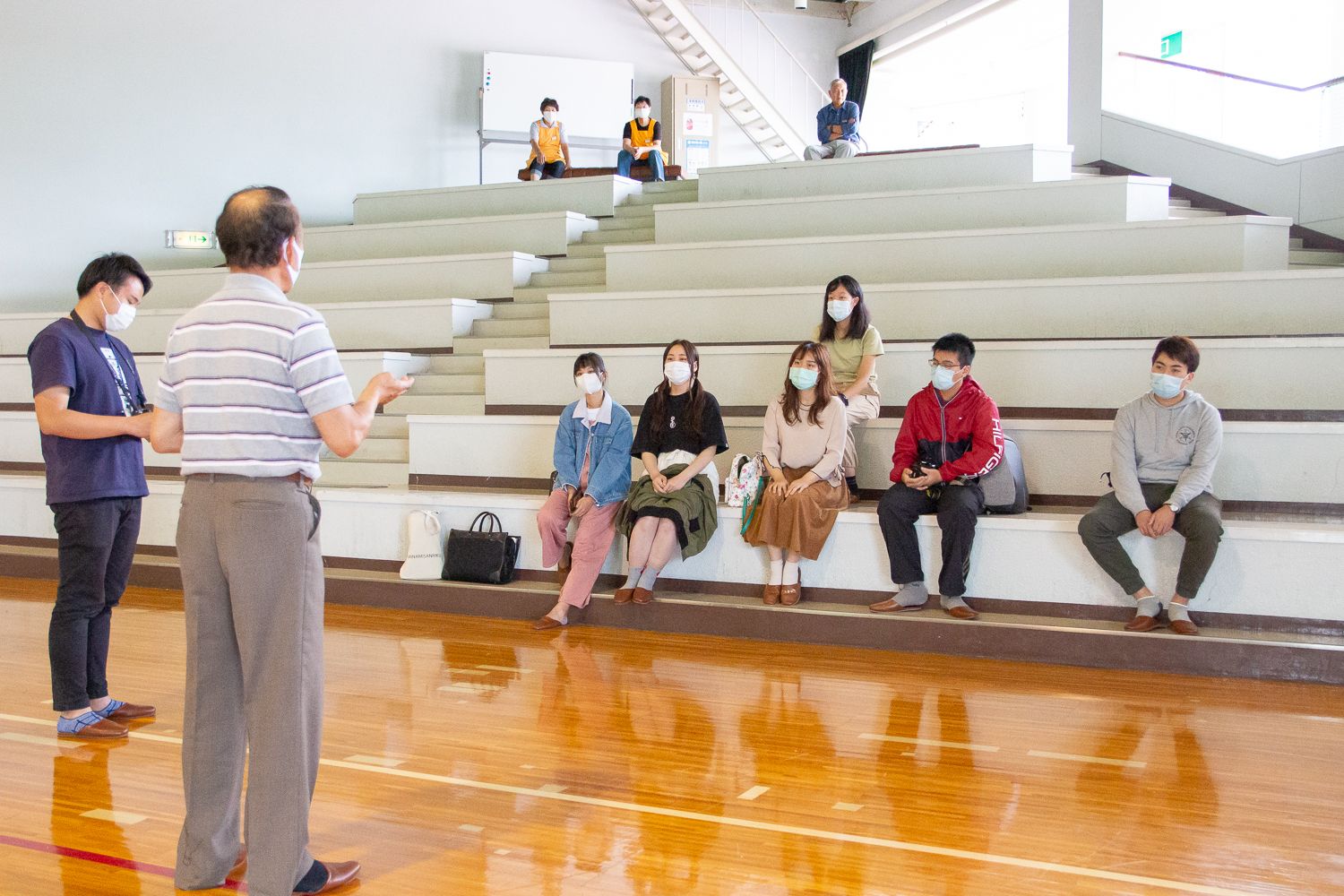 体育館の観客席に間隔をあけて座ったマスク姿の男女が、床に立つ村岡賢一保存会長から説明を受けている様子で、さらに上段には数名が離れて座って話を聞いている写真