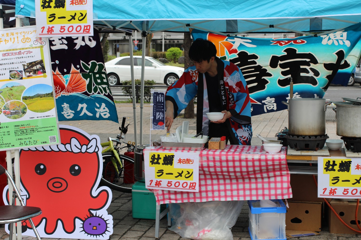 青いテントの下の屋台には大きな鍋が並び、「本日限り牡蠣ラーメン1杯500円」と書かれているポップが貼られた前で、法被を着た男性が牡蠣ラーメンに具をよそっている写真