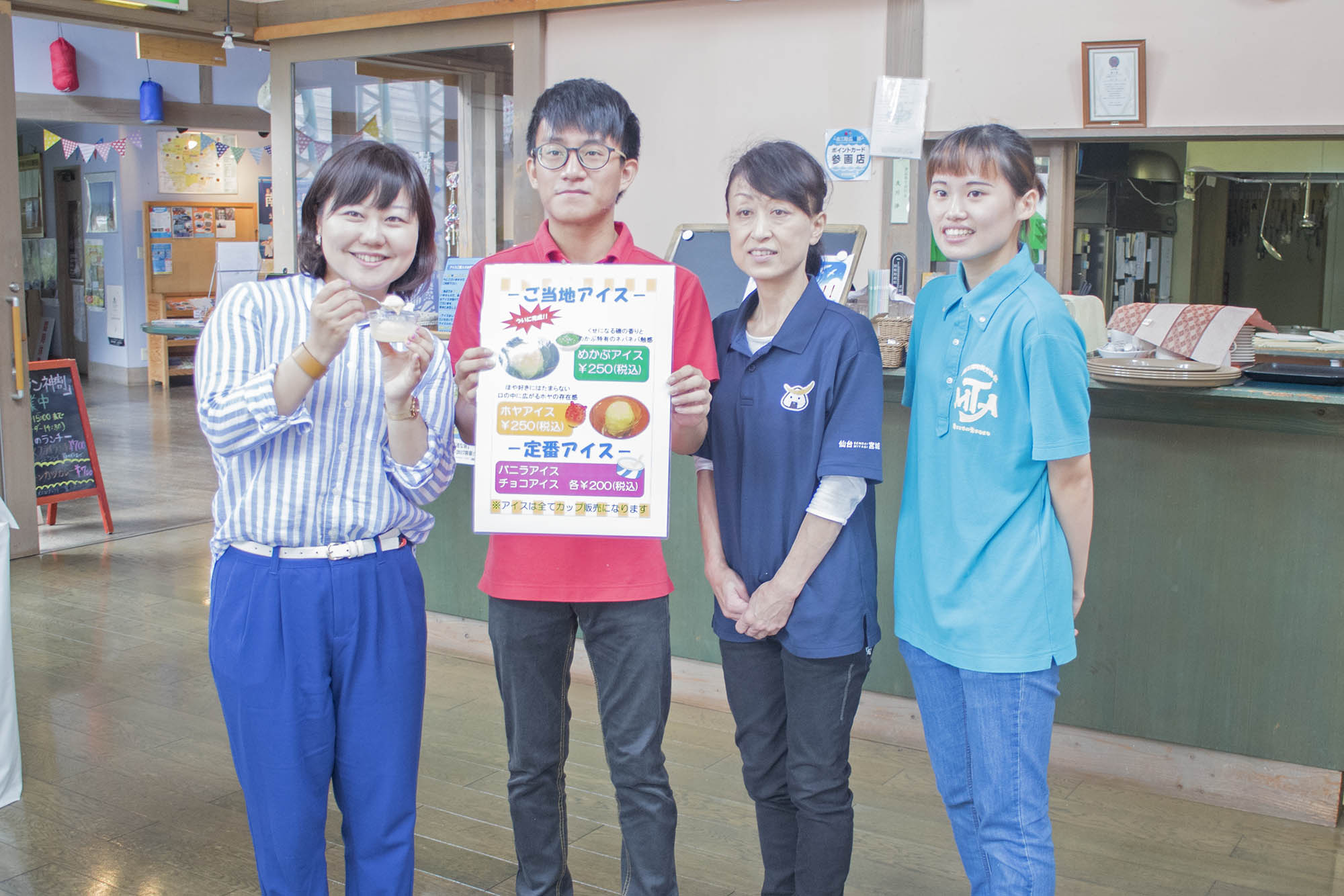 レストランで、リポーターの女性がご当地アイスを手にしながら、アイスの案内看板を持つ男性と、ネイビーと水色のポロシャツを着た女性2人と一緒に並んでいる写真