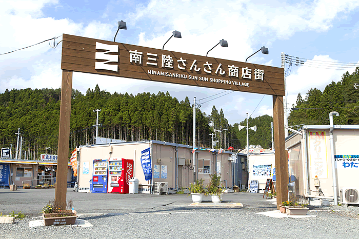 青空が広がり、周囲を山や木々に囲まれた背景には仮設のような平屋の商店が並んでおり、手前に「南三陸さんさん商店街」と書かれた木製のアーチ型の看板が設置された商店街の入口の写真