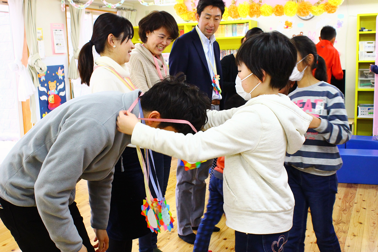 笑顔の先生が4人ほど並んで立っており、手前の先生が児童館の利用者である子どもにカラフルな手作りの首飾りをかけてもらっている様子の写真