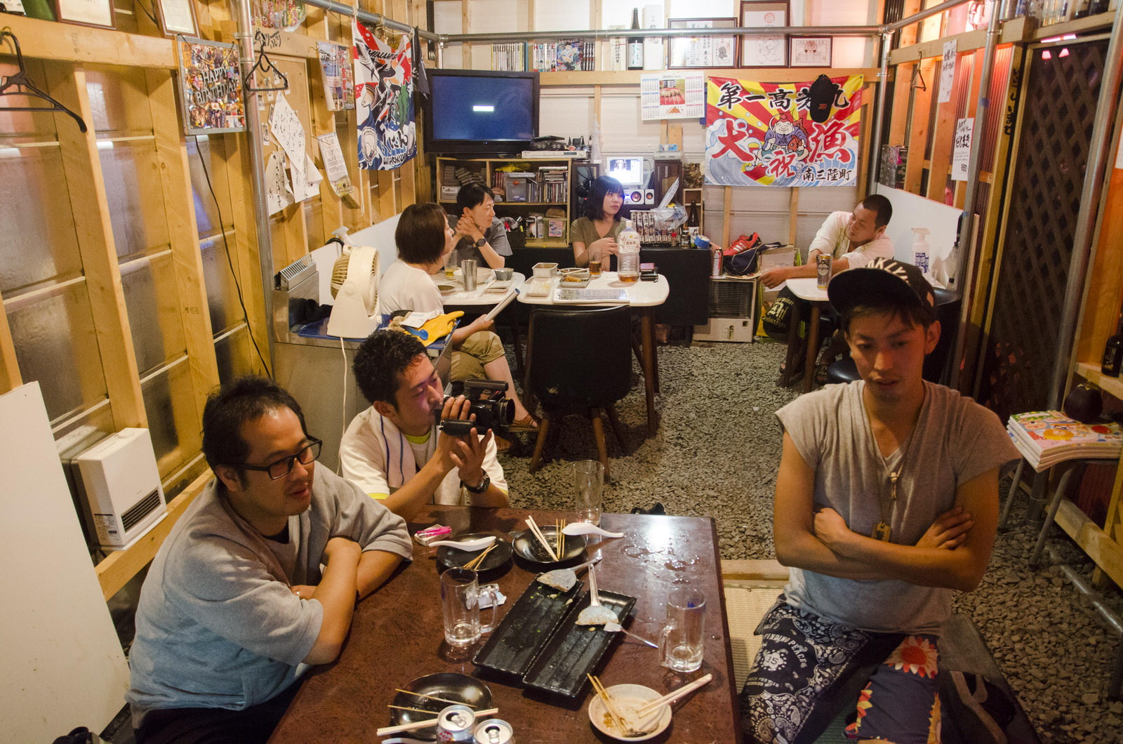 木造の内装が見えるカジュアルな店内の奥のテーブル席で複数の客が談笑しながら飲食を楽しんでおり、手前のテーブルで腕を組んだ井尻さんが真面目な顔をしてカメラを構えた男性と眼鏡をかけた男性とテーブルを囲んで座っている写真