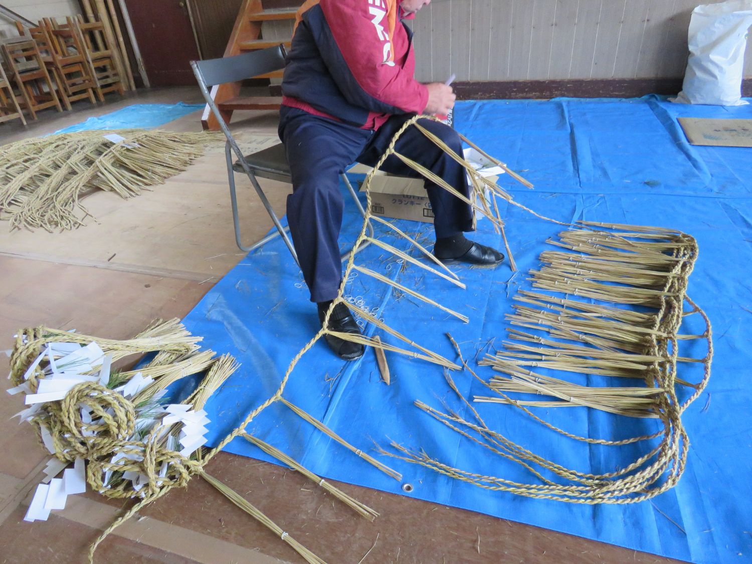 ブルーシートの上に15下げ一間半の長さのしめ縄が重ねて置かれ、パイプ椅子に座った山内貞行さんがしめ縄を作る様子の写真
