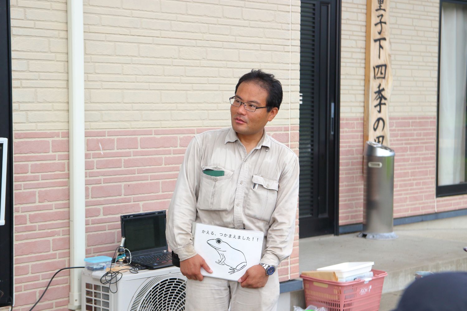 ベージュ色の作業着を着た男性講師が、カエルの絵と「かえる、つかまえました！！」という文字が書かれた紙を持って参加者に説明している様子の写真