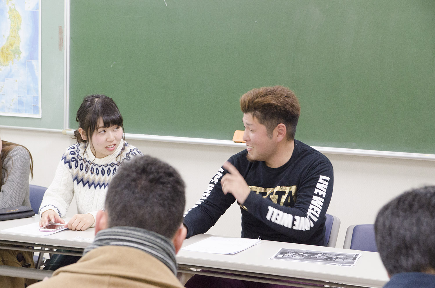 教室内で黒板を背に長机の席に座った千葉さんが隣の女性に指をさしながら話しかけ、周囲に数人の人物がいる写真