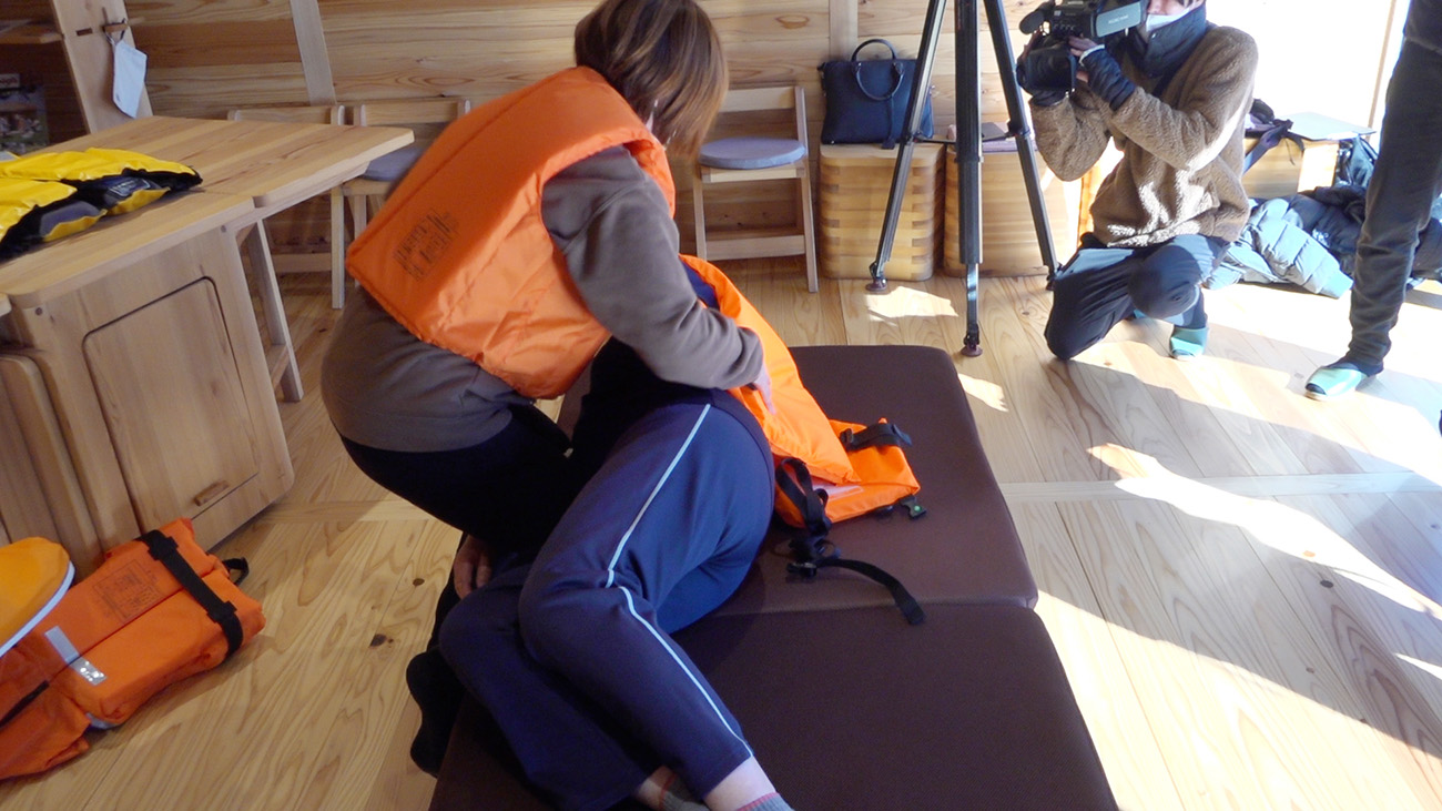 オレンジ色のライフジャケットを着た人がマットの上に横たわり、もう一人がその人を支えていている様子の写真