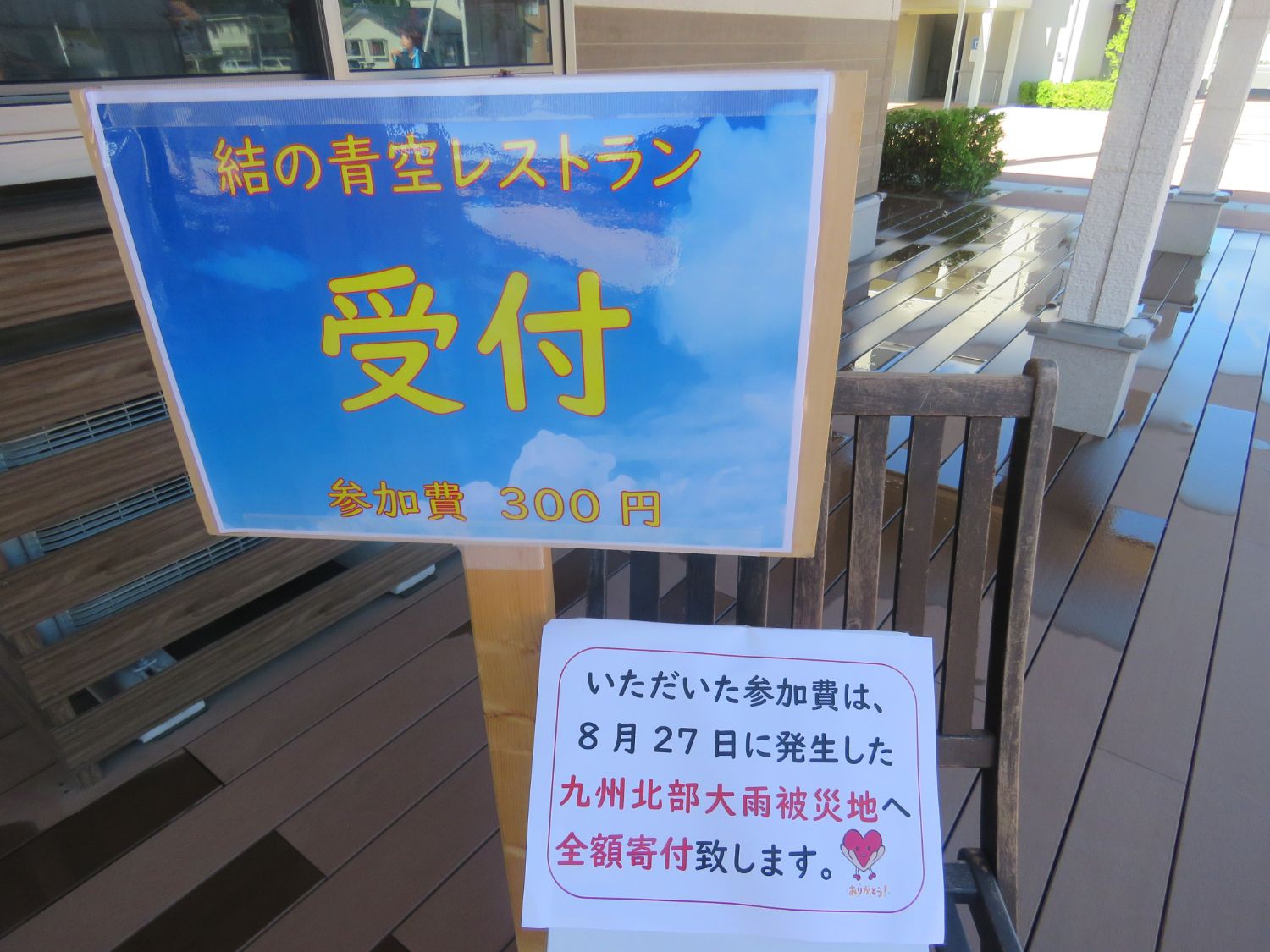 木製の椅子の前に「結の青空レストラン 受付 参加費300円」と書かれた青空を背景にした案内板が立てられ、その下には「いただいた参加費は、8月27日に発生した九州北部大雨被災地へ全額寄付致します」と赤字と黒字で書かれたメッセージが添えられている写真。