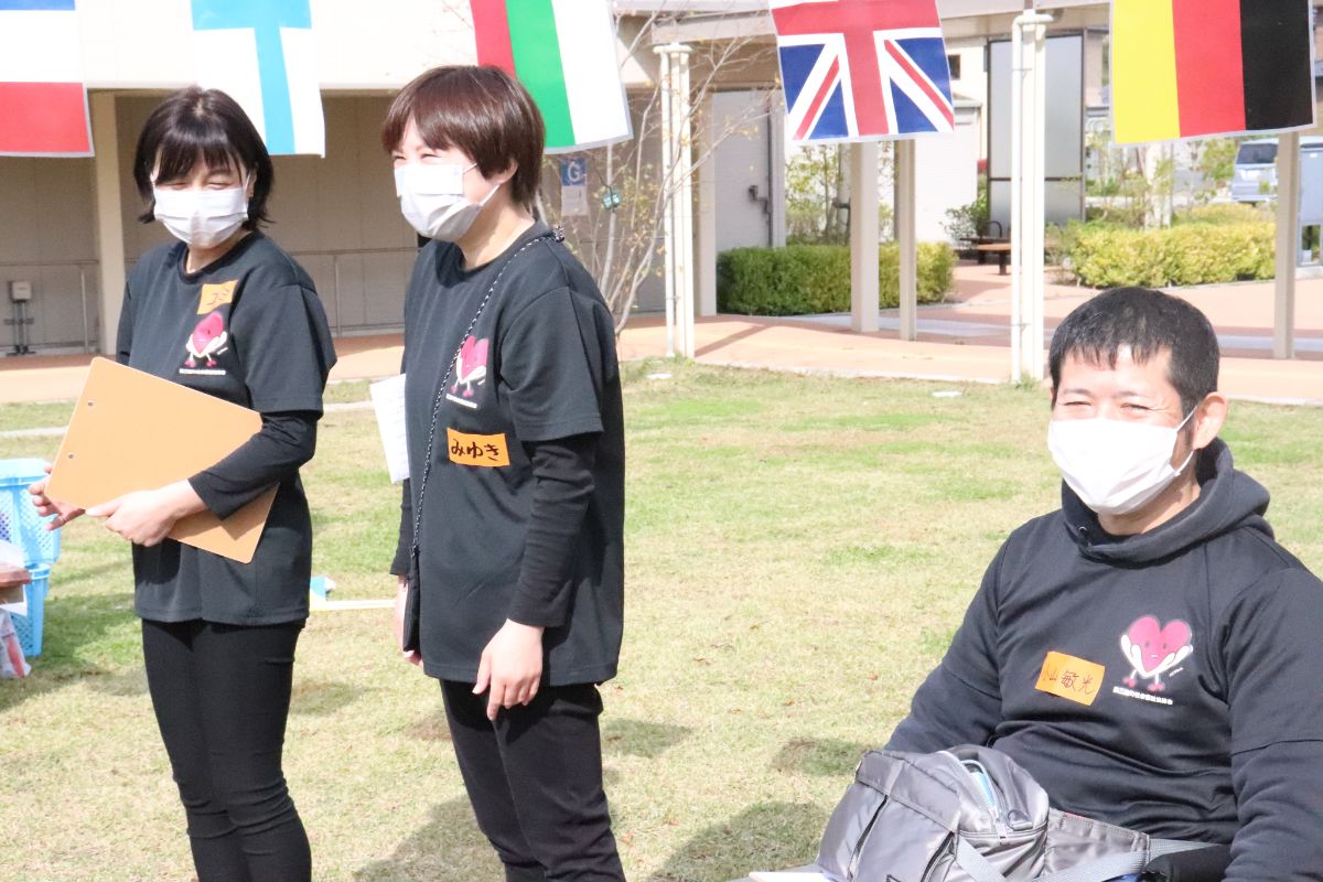 万国旗が掲げられた屋外で、黒いスタッフTシャツを着でマスクをつけた2人の女性が立ち、1人の男性が座っている様子の写真