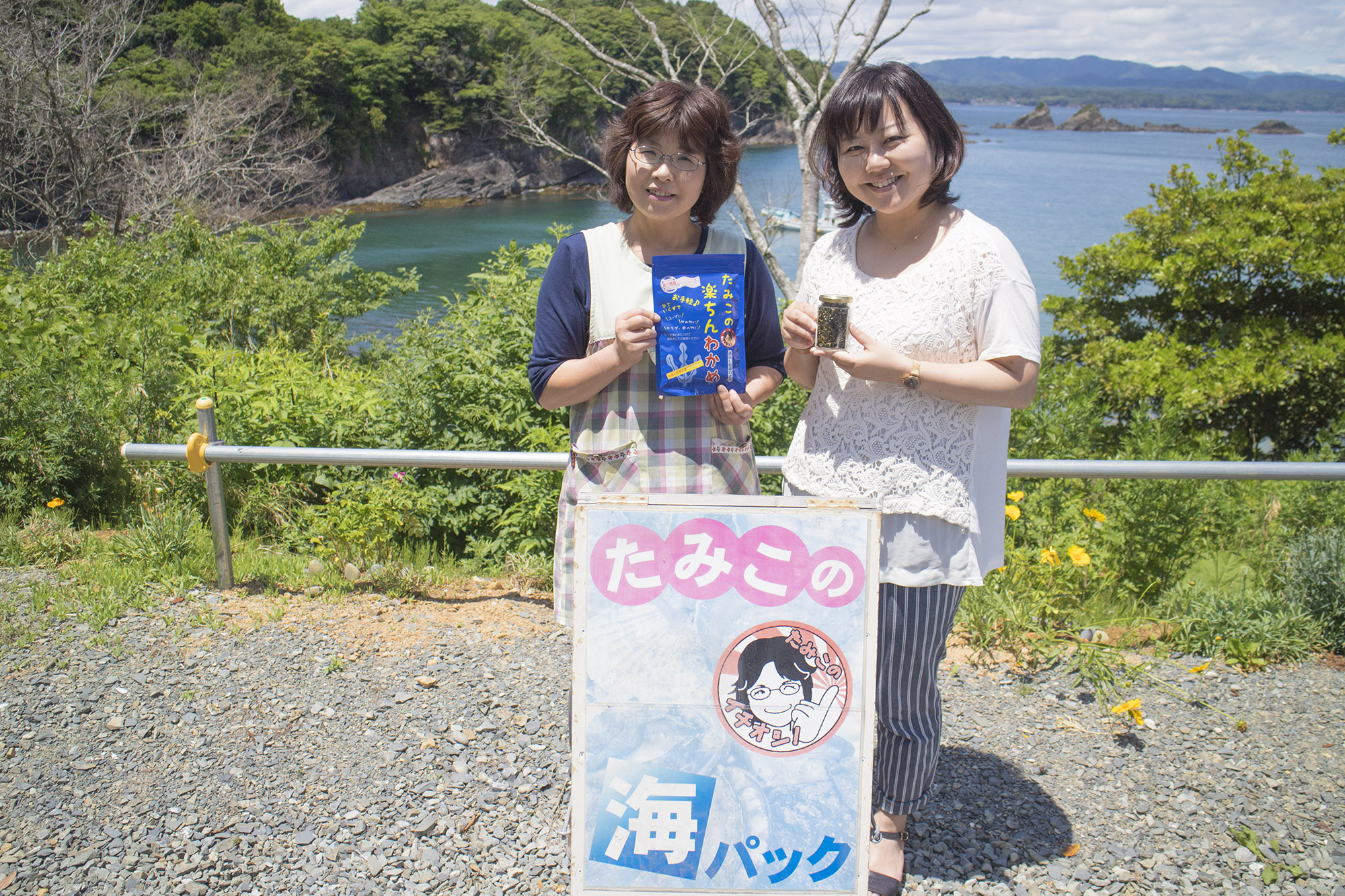 自然の緑と海を背景にしてエプロン姿の阿部さんとリポーターの女性が「たみこの海パック」と書かれた看板の後ろで加工された海藻商品のパッケージを手に笑顔で立っている写真
