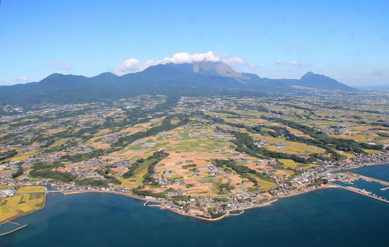 雲をかぶった山並みのふもとに、住宅地、田畑が広がり海岸線まで広がっている様子が見える航空写真