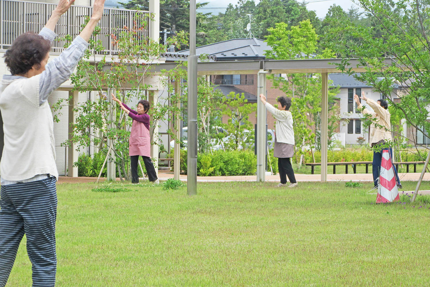 芝生の広場で輪になって広がった複数の高齢女性が、手を上げたり伸ばしたりする体操をしている写真