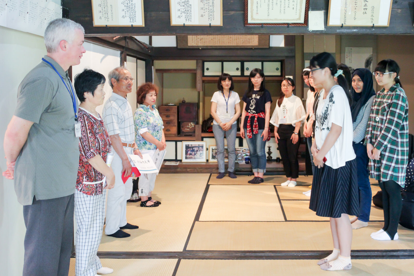 和風の家の中で地元の年配の人たち4人が並び、向かい合って立つ白いシャツを着た代表の女性が丁寧に挨拶をしている対面の場面の写真