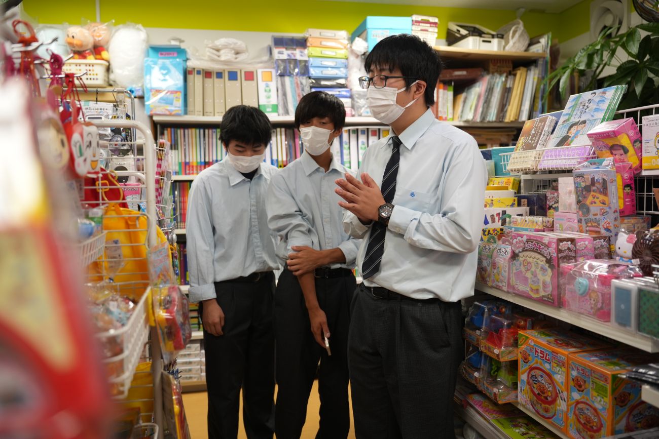 文房具やおもちゃが並ぶ店内で、男子高校生2人と男性が会話をしている様子の写真