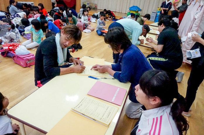 大勢の子どもたちが広い部屋に集まり、脇に置かれた机では保護者と教師などが書類の記入をしている様子の写真