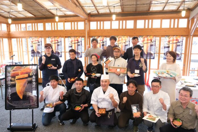 木製の梁がある明るい室内で、料理人やスタッフ15人が料理や食材を持ち、笑顔で片手を握りこぶしにしてポーズを取っている、試食会の様子の写真