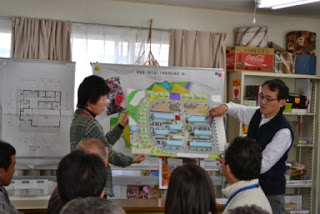 前に立つ男性が関係者の方とまちづくりの図面を参加者に見えるように掲げて見せ、参加者に向けて地域計画の説明を行っている様子の写真