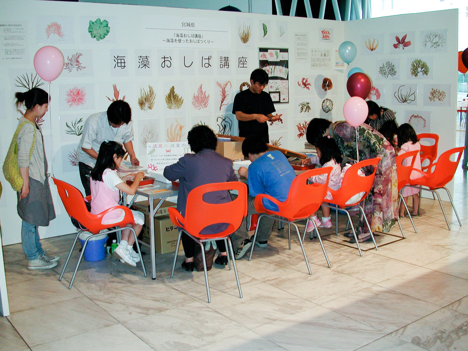 様々な海藻の押し葉作品が展示されている「海藻おしば講座」と書かれた壁の前で、オレンジ色の椅子に座った複数の子どもと大人が参加している様子の写真