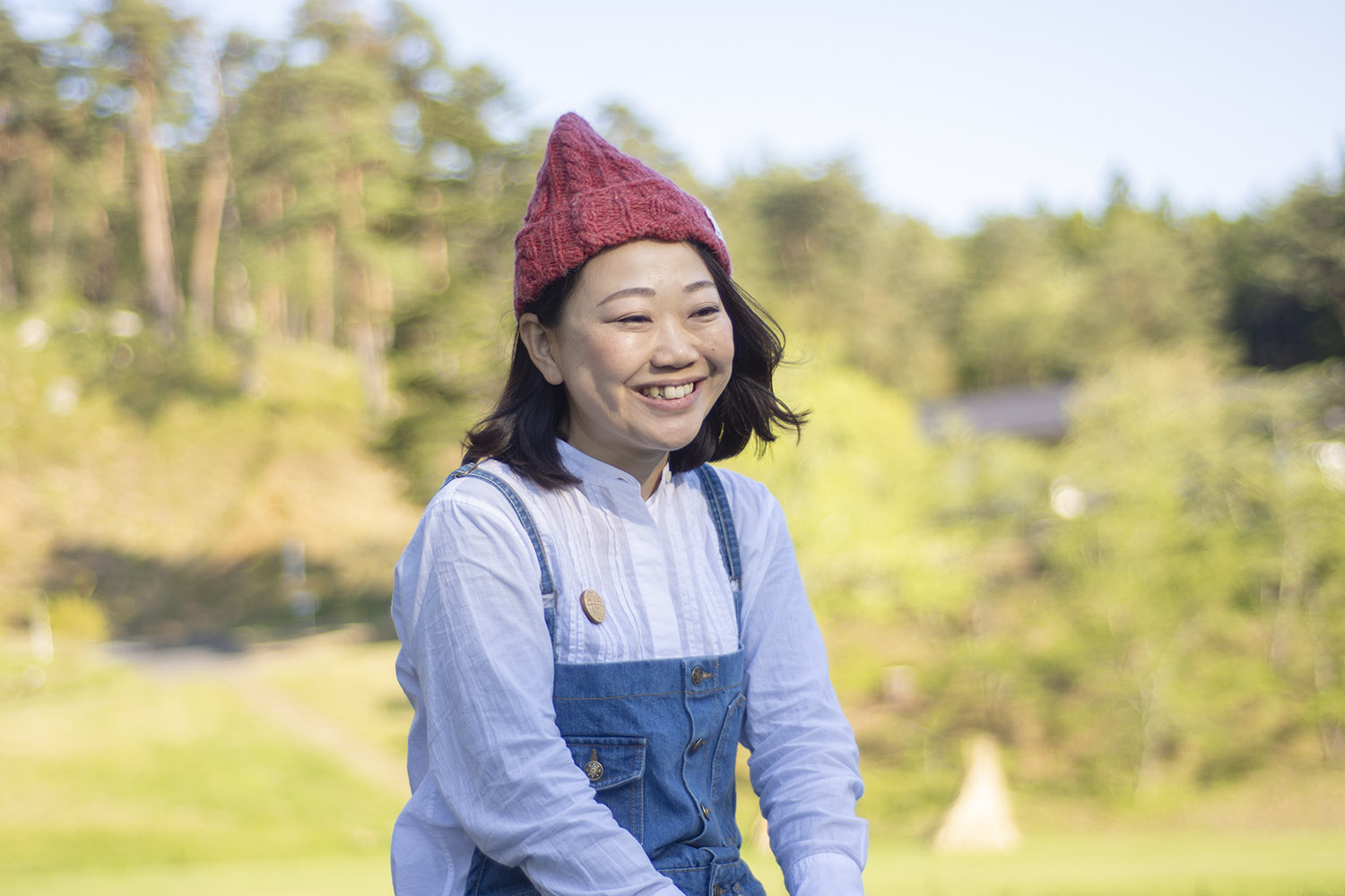 赤いニット帽をかぶり白いシャツとデニムのオーバーオールを着た女性が、屋外で微笑んでいる表情をアップで撮影した写真