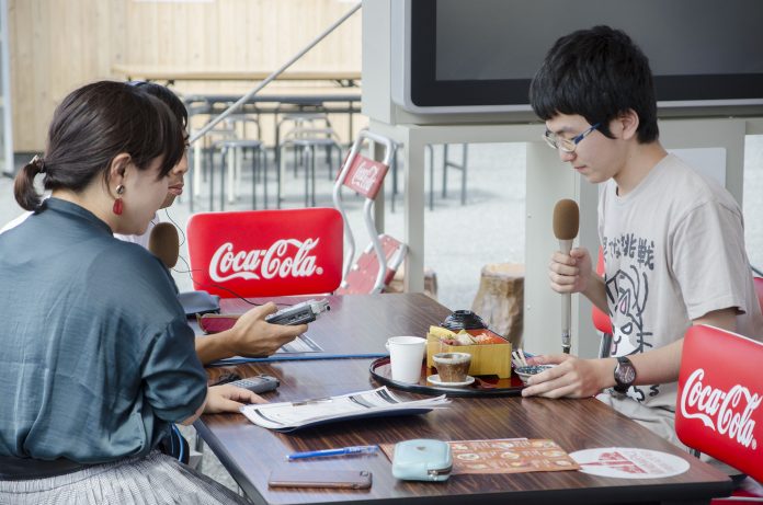 女性リポーターがと録音機を持った男性が、長机の対面に、海鮮丼を前にして座っている眼鏡をかけた平野さんにインタビューしている様子の写真