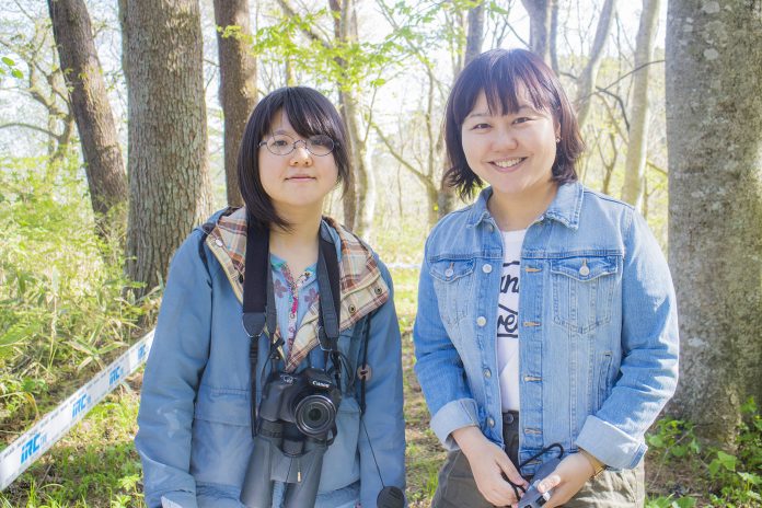 森林の中で首にカメラや双眼鏡をかけた大渕さんと女性リポーターが並んで立っている写真