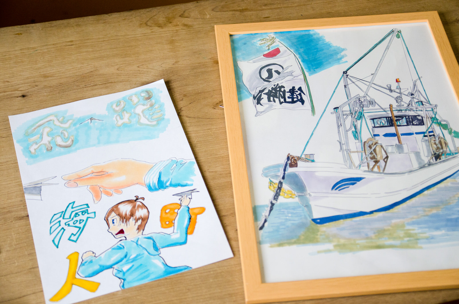 紙飛行機を投げている若者の絵と、額に入った漁船の絵が並んで置かれている写真