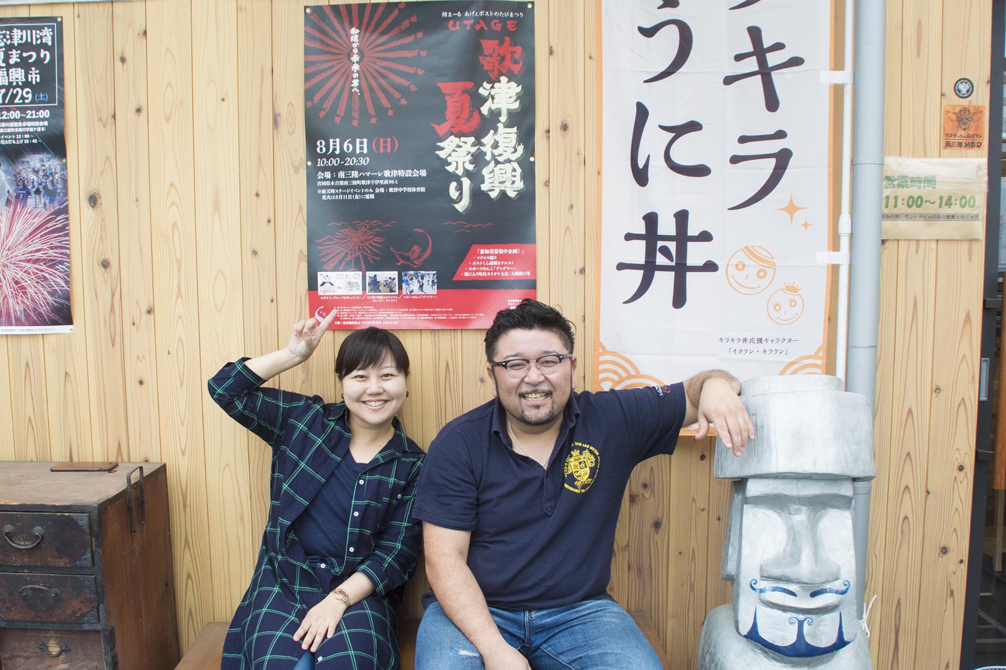 木目の外壁を背に、左側に座る女性リポーターが壁に貼られた歌津復興夏祭りのポスターを指さし、右側にいる紺色のポロシャツ姿の男性が隣にあるシルバーのモアイ像にひじをかけて笑顔を見せている写真