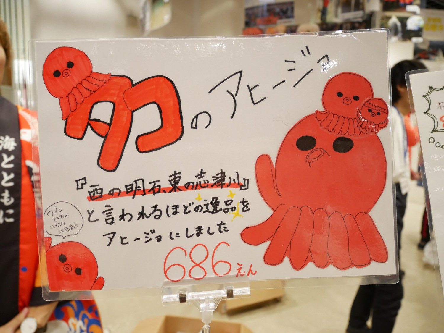 赤いタコのキャラクターが大きく描かれたポップ広告で、「西の明石、東の志津川」と言われるほどの良質なタコを使用したアヒージョを紹介しており、価格は686円と手書きで表示されている写真