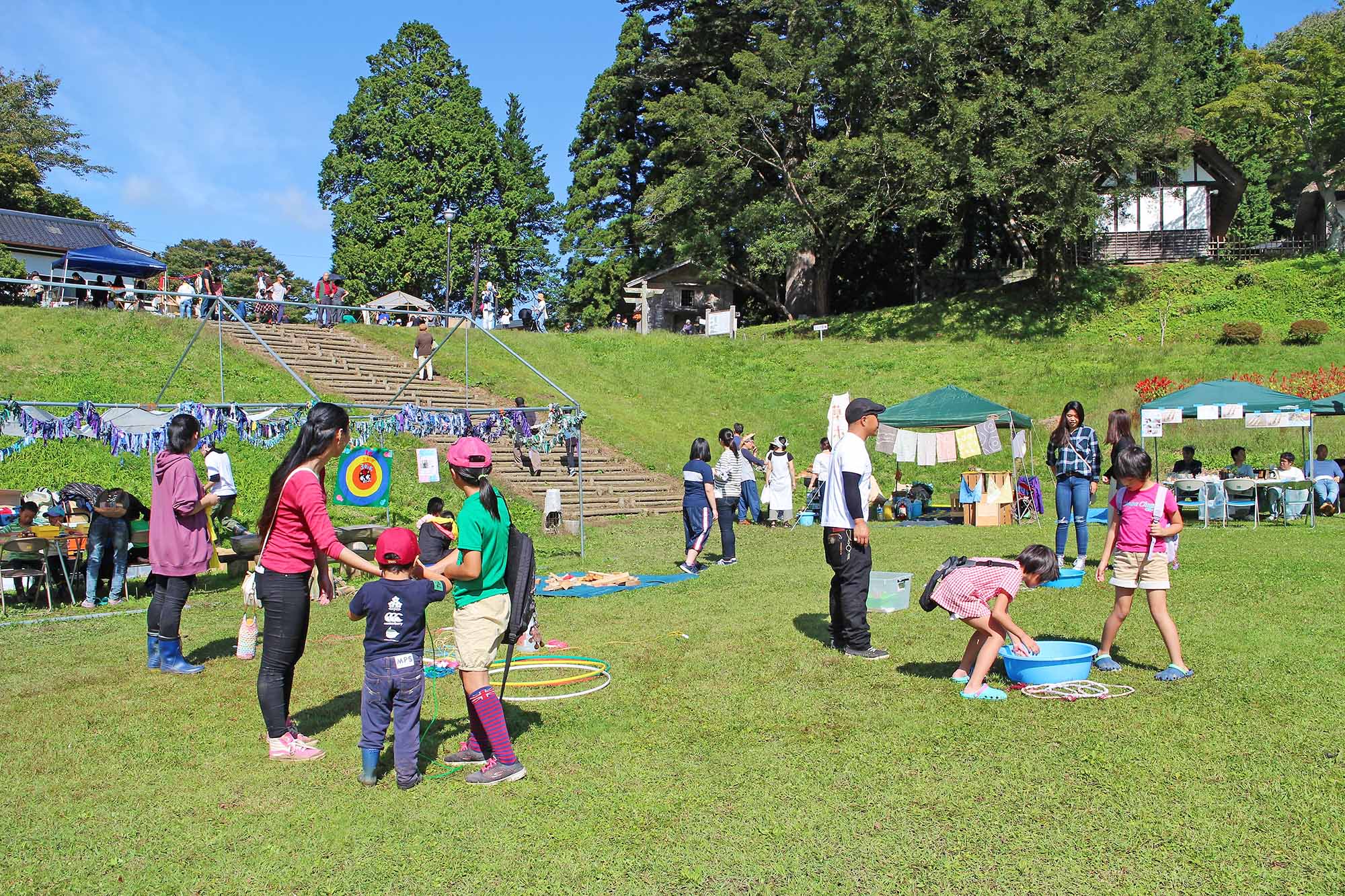 子どもたちと大人が芝生の上で輪投げや水遊びをしながら楽しんでいる、屋外イベントの様子の写真