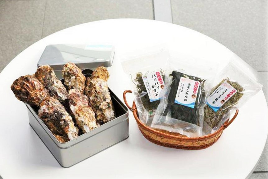 白いテーブルの上に、缶に入った殻付き牡蠣と、乾燥わかめや昆布などが袋詰めで籠に入って並べられている写真