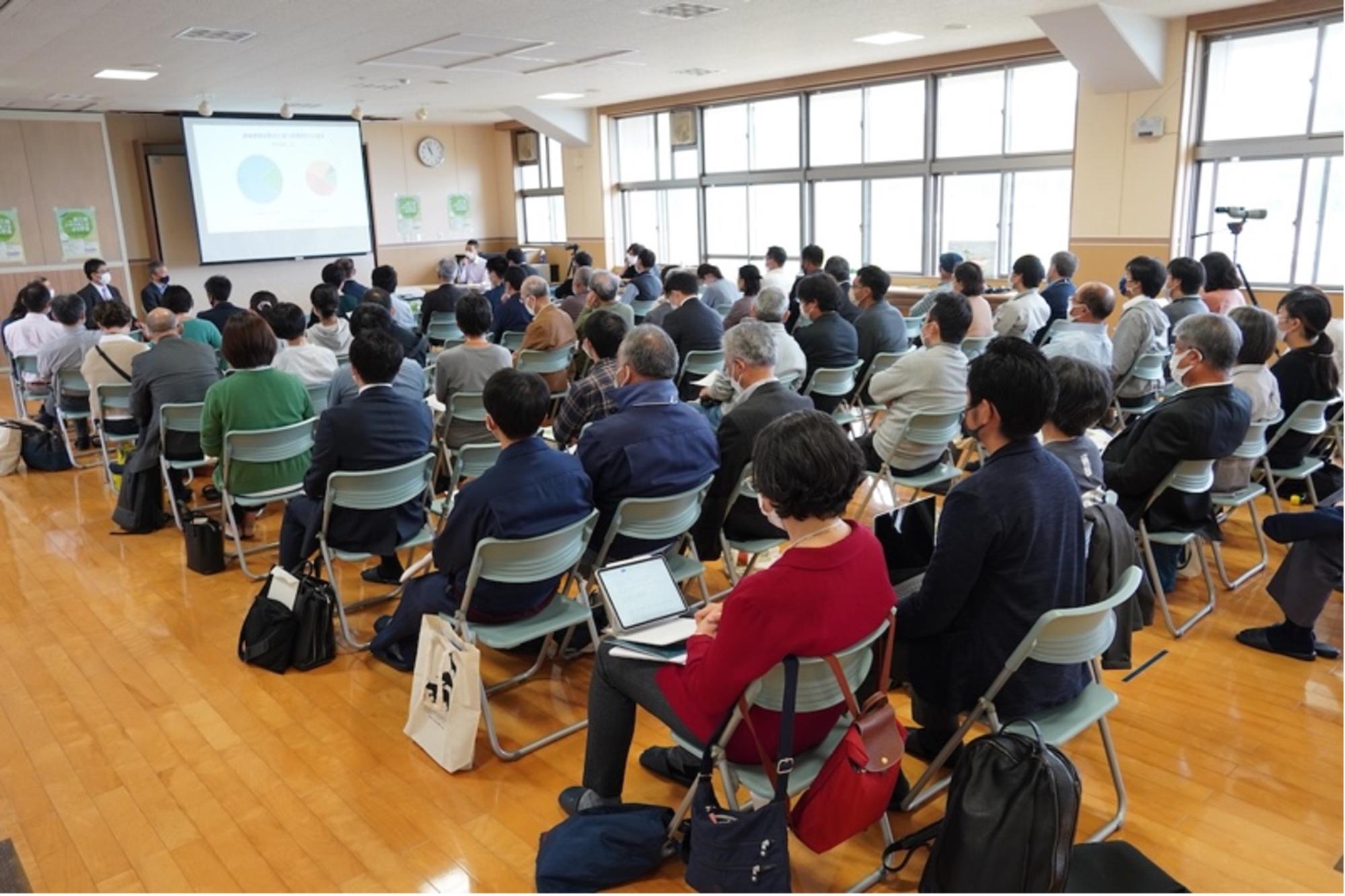 椅子に座った多くの参加者たちが前方のスクリーンに注目している様子を後ろから撮影した写真