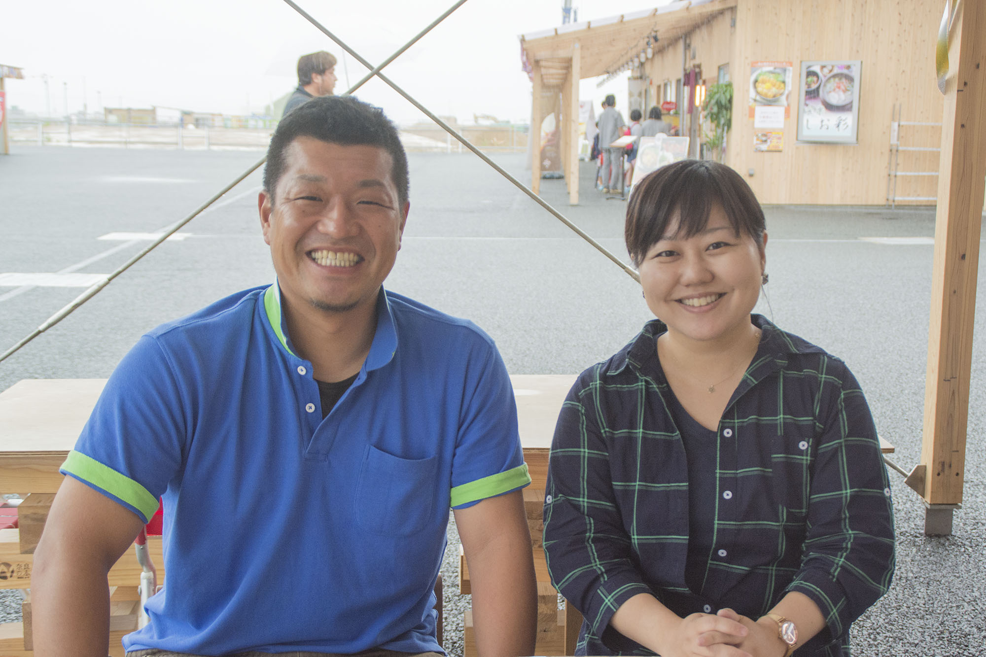 青いポロシャツを着た工藤さんとリポーターの女性が、商店街の店舗を背景に木製のベンチに並んで座ってにこやかにこちらに向かって笑っている写真