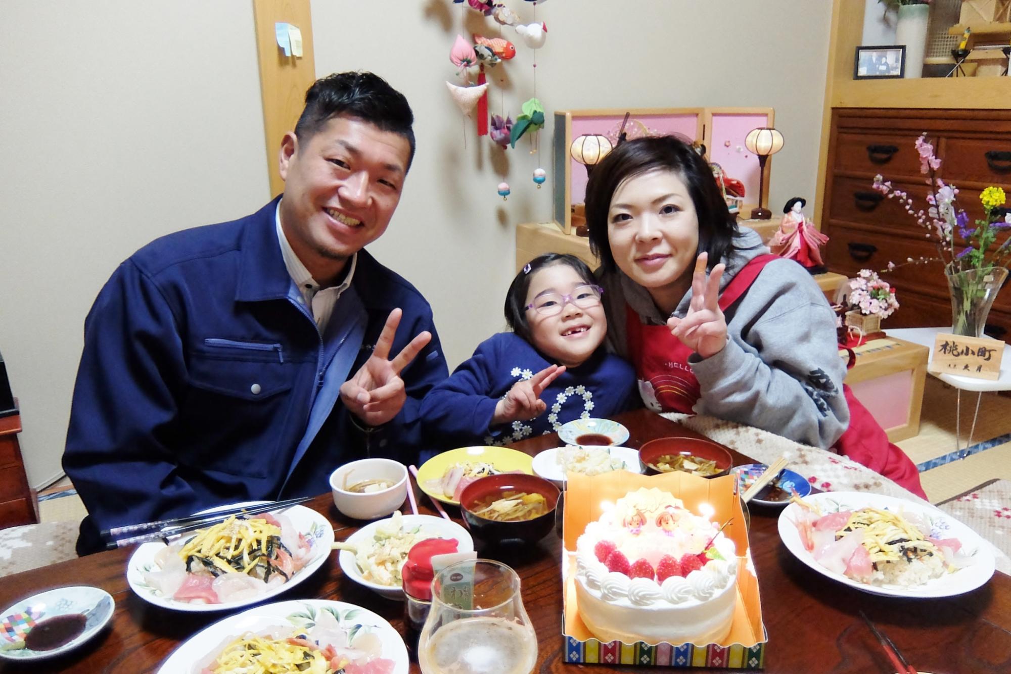 和室で工藤さん夫婦と光奈乃ちゃんが雛飾りのある背景の前で、料理とケーキが並ぶ食卓を囲んで笑顔でピースサインをする写真