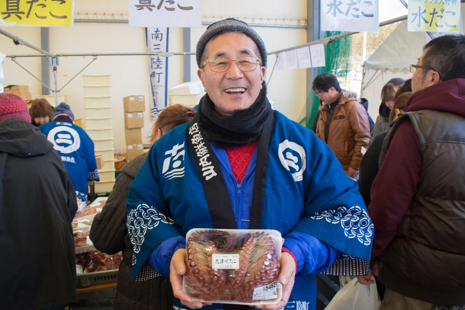 屋内の市場の一角で、青い法被を着た年配の男性が笑顔で正面を向きながら両手でパック詰めされた大きな茹でだこを持ち写っている写真