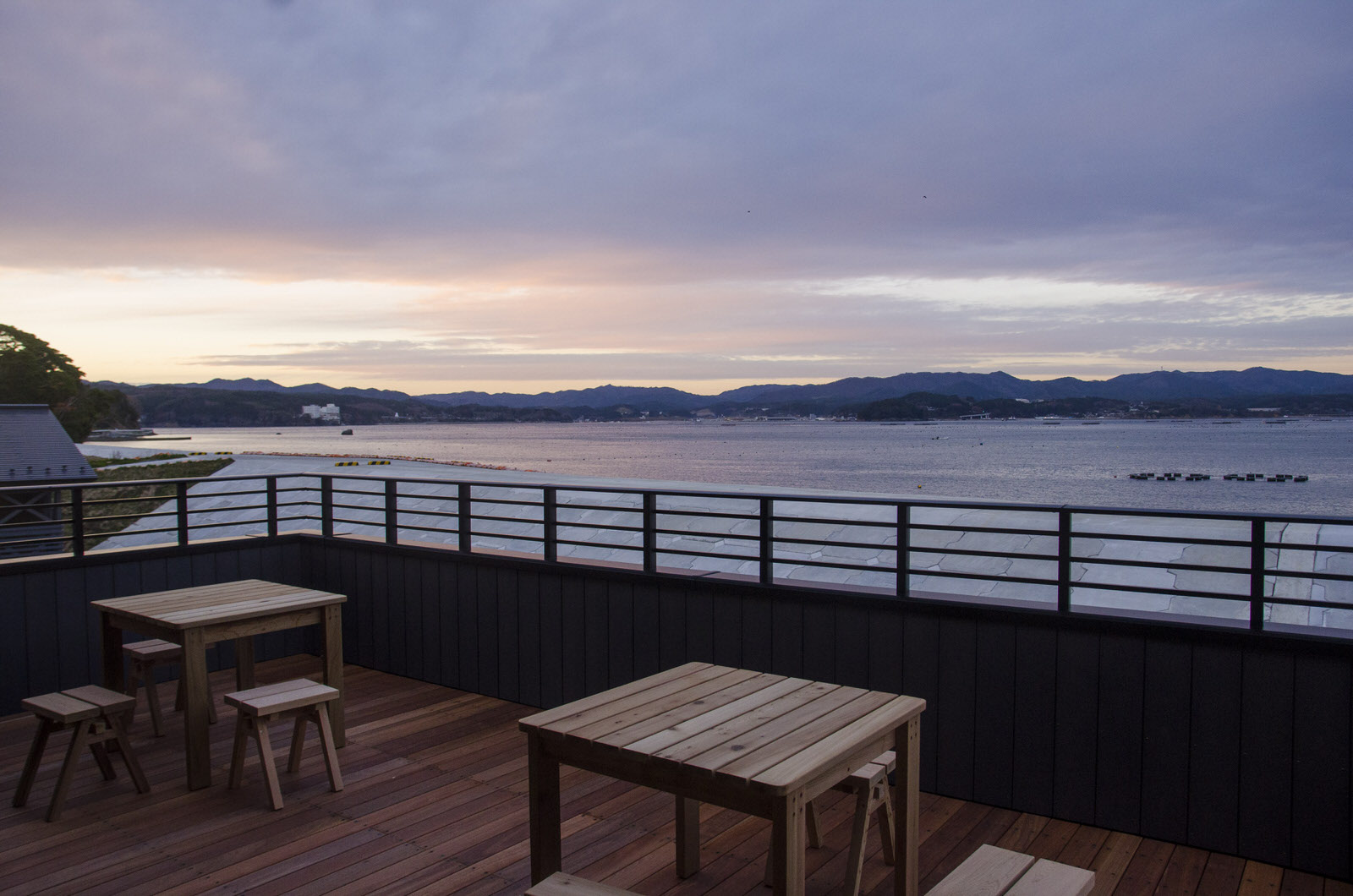 海と山が望める夕暮れ時の2階のテラス席に、木製のテーブルと椅子が置かれたウッドデッキの写真
