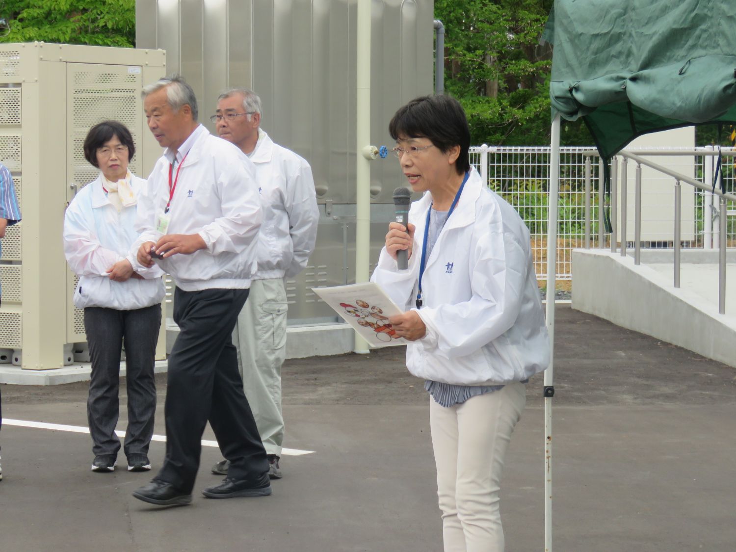 お揃いの白いジャンパーを着た数人の高齢者が立って見守っているなか、左手にクリアファイル、右手にマイクを持った女性が話している写真