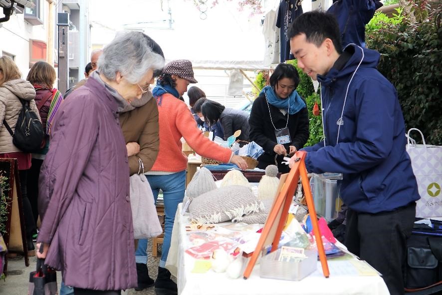 編み物や帽子などの手工芸品が並ぶ屋台で、男性の出店者が笑顔で年配の女性に対応している手作り品の販売風景の写真
