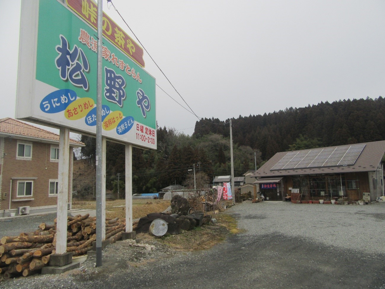 「松野や」という名前の飲食店の看板があり、看板には「峠の茶や」「農家手づくりうどん」「うにめし」「あさりめし」「ほたてめし」などの文字が書かれ、背景には木々に囲まれた建物が見える様子が写っている写真