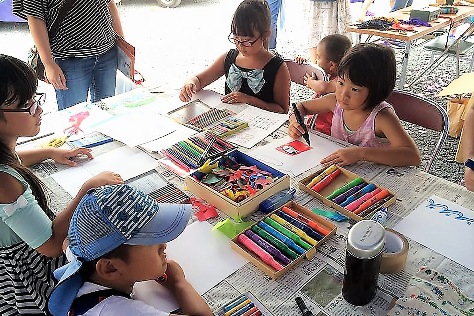数人の子どもたちが屋外の机で色鉛筆やクレヨンを使って自由に絵を描いている様子で、机の上には色ペンがたくさん並んでいる写真