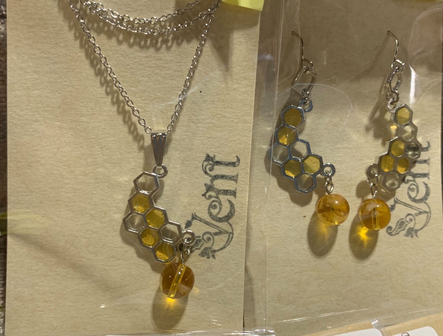 ハチの巣を模した六角形をつないだ金属パーツに、黄色いガラス玉がついたネックレスとイヤリングのセットがパッケージに入って並べられている写真