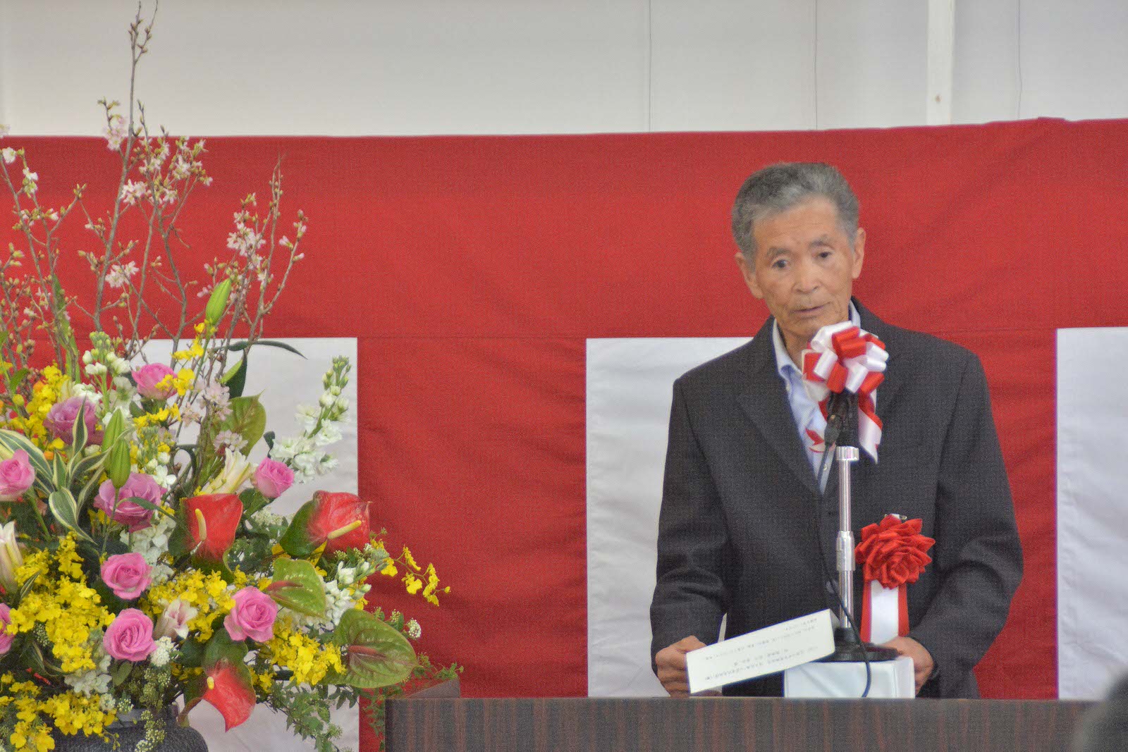 紅白幕を背に、豪華な花が飾られた演台に立ち、マイクに向かって話をする宮川さんの写真
