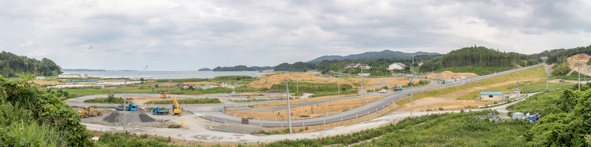 海に面した丘陵地の復興の定点カメラによる風景でグレーがかった曇空の中、左奥には海が広がり中央にはアスファルト舗装済みの道路が縦横に伸びており、すでに車両が走行し道路の間には造成中の更地が広がり、数台のショベルカーやダンプトラックが工事を進めている様子を写した写真